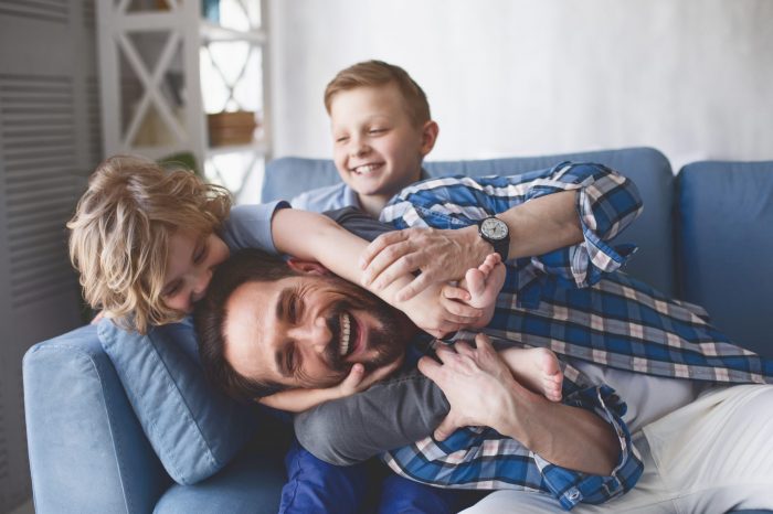 Portrait of smiling father and outgoing sons having fun on cozy couch in apartment